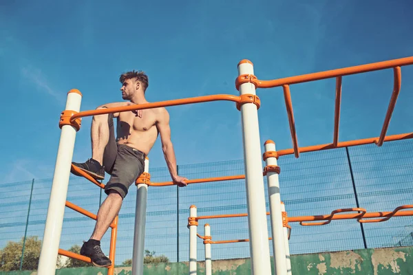 Sportman met kale romp monkey bars bovenop. Man atleet klimmen op metalen jungle gym op blauwe hemel. Macho ontspannen na training buiten. Sport, gezondheid en energie concept — Stockfoto