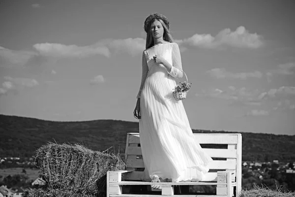 Chica albina con flores, belleza natural. Mujer novia en vestido de novia en banco de madera. Mujer sensual en guirnalda sobre pelo largo y rubio. Modelo de moda en el soleado cielo azul de vacaciones — Foto de Stock