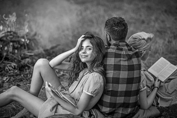 Camping in wilderness. Turn back guy looking at fire while two beautiful girls read book. Smiling brunette with braces leaning on her friend — Stock Photo, Image