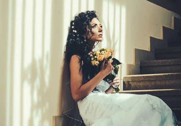 Чуттєва жінка з весільним букетом. Жінка з квітами сидить на сходах. Дівчина з весільним макіяжем і зачіскою. Наречена в моді біла сукня. Весілля і очікування на майбутнє — стокове фото