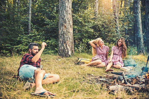 Férias de verão, fim de semana na floresta, camping. Duas raparigas sentadas na relva enquanto o tipo atira paus para o fogo. Conceito de natureza e ambiente — Fotografia de Stock