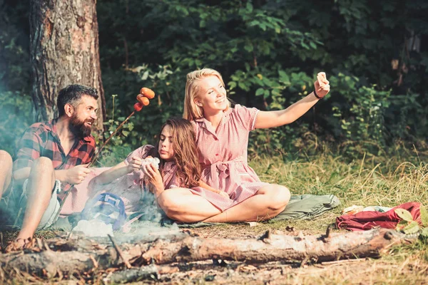 Женщина делает фото или видео со смартфоном в лесу. Друзья делают селфи с сосисками у костра. Бойфренд и подружки улыбаются для мобильного телефона. Новые технологии и современная жизнь, винтаж — стоковое фото