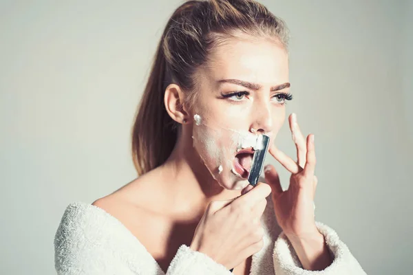 Barber and shaving concept. Woman with face covered with foam holds straight razor in hand. Girl on busy face wears bathrobe, grey background. Lady shaves her face with sharp blade of straight razor. — Stock Photo, Image