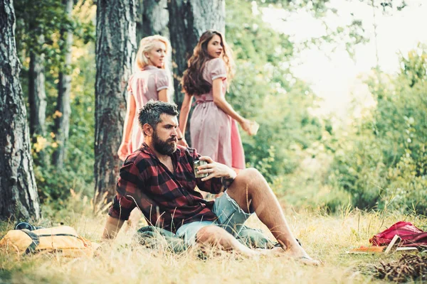 Kemping w słonecznej łące. Dwie dziewczyny, patrząc wstecz na człowiek siedzi na trawie. Brutalny facet pije kawę przy ognisku — Zdjęcie stockowe