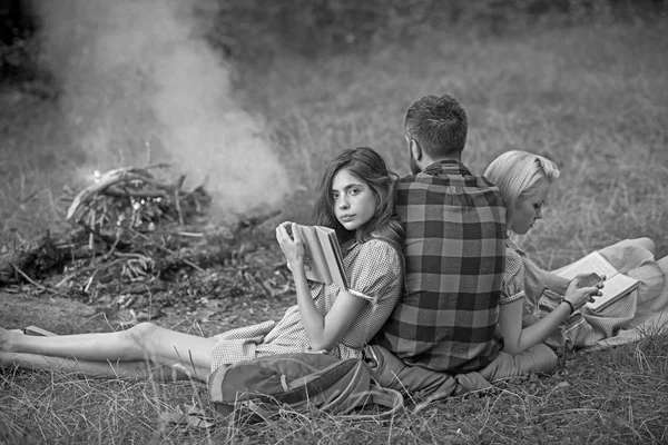 Vahşi doğada kamp. İki güzel kız kitap, sürdürülebilir eğitim kavramı okurken ateş arayan adam geri çevirmek — Stok fotoğraf