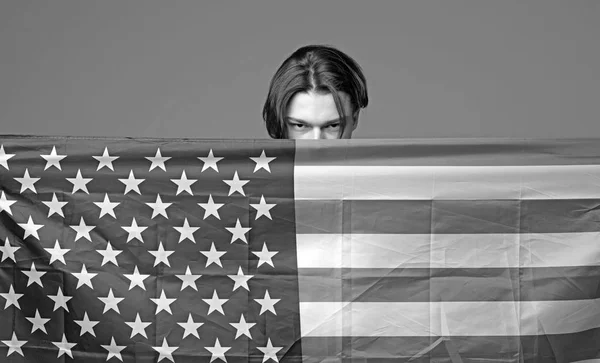 Mann mit ernstem Gesicht hält US-Fahne in der Hand und versteckt sich hinter Fahne. Typ versteckt sich in den USA, Amerika beschützt ihn, grauer Hintergrund. Privatsphäre und Verschwörungstheorie. — Stockfoto