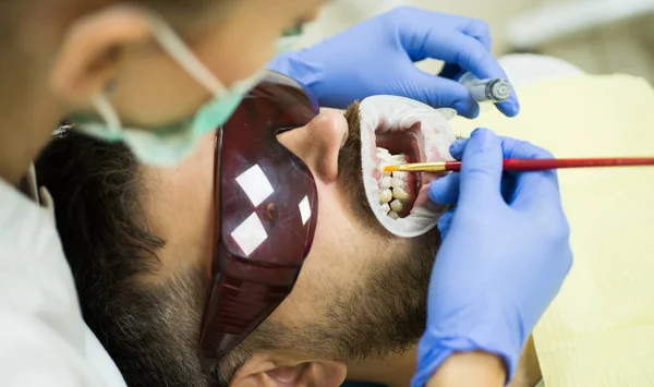 Dentiste examinant les dents des patients à la clinique. Homme barbu ayant des dents examinées chez les dentistes. Jeune homme souriant après la visite dentiste sur fond flou lumineux . — Photo