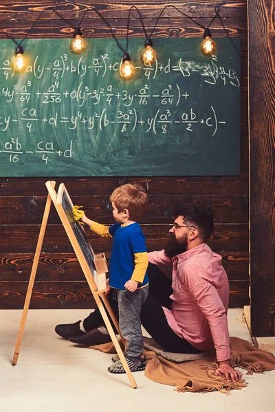 Pequeño genio aprendiendo matemáticas. Profesor o padre ayudando al niño a resolver la ecuación en pizarra. Tipo barbudo con camisa rosa sentado en el suelo — Foto de Stock