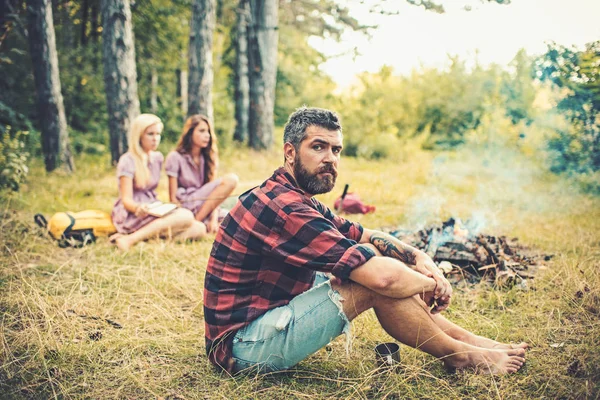 Бородатый мужчина расслабляется у костра с женщинами на размытом фоне. Хипстер с бородой сидит у костра. Друзья любят ходить в походы в лесу. Летние каникулы. Кемпинг, туризм и путешествия — стоковое фото