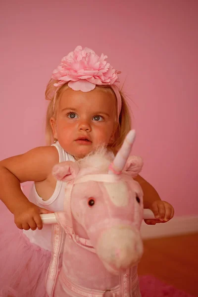 Estar numa sela. Criança pequena usar faixa de cabelo. Acessório de cabelo. Pequena menina loira montar brinquedo cavalo. Menina com penteado longo. Penteado infantil para meninas. És tão bonita, pequenina. — Fotografia de Stock