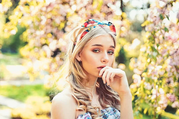 Chica en la cara pensativa de ensueño, tierna rubia cerca de flores de sakura, fondo de la naturaleza. Concepto de primavera. Lady camina en el parque el soleado día de primavera. Mujer joven con chal en la cabeza disfrutar de la naturaleza — Foto de Stock