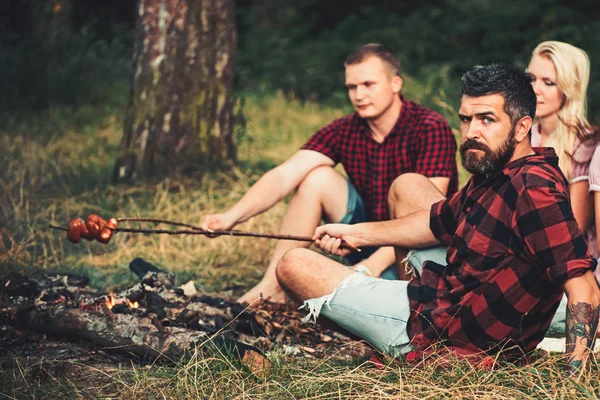 友達と男ロースト ソーセージひげを生やした。男性と女性の森でたき火でピクニックがあります。人々 は火を料理します。彼氏や恋人は、夏のキャンプをお楽しみください。調理食品および休暇を旅行します。 — ストック写真