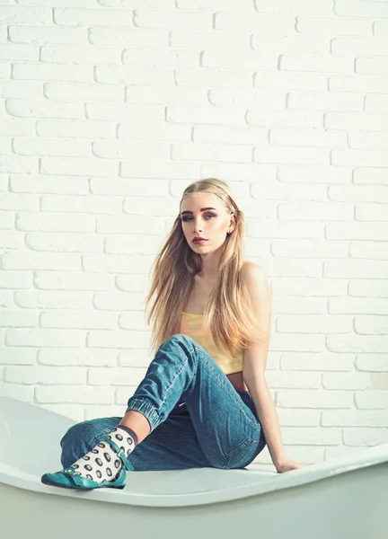 Blond model with messy hair sitting on the edge of bath tube in front of white brick wall