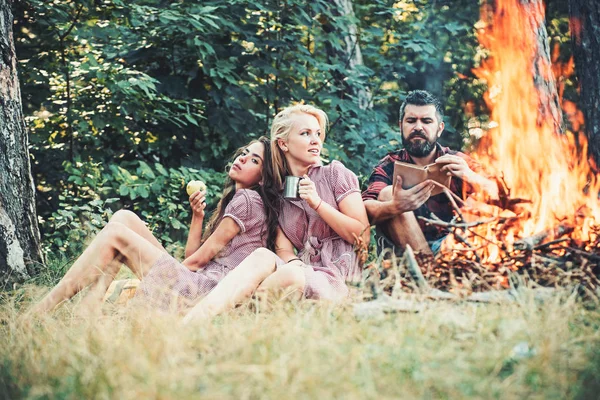 Gruppe von Freunden campiert im Wald. Mann liest Buch von Feuer. schönes blondes Mädchen trinkt Tee aus Becher, während ihre Freundin grünen Apfel isst — Stockfoto