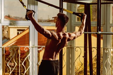 Spor ve spor salonu konsept. Adam çıplak gövde ve geri içinde jimnastik salonu kas eğitim, trx tadını çıkarın. Gövde, sporcu, atlet, kas maço mu egzersiz trx döngüler ile pencere arka plan üzerinde olan adam.