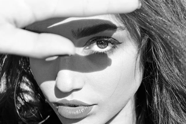 Woman on calm face with makeup, covers sunlight with hand, defocused. Lady looks attractive, close up. Bright sunlight concept. Girl brunette with red lips on sunny day.