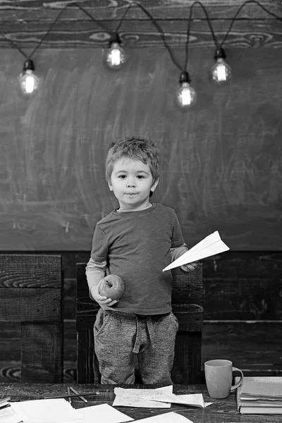 Μικρό αγόρι κρατώντας χαρτί αεροπλάνο και η apple στα χέρια του. Μαθητής στο μπλε T-shirt στέκεται πίσω από το τραπέζι. Εκπαιδευτική έννοια παιχνίδι — Φωτογραφία Αρχείου