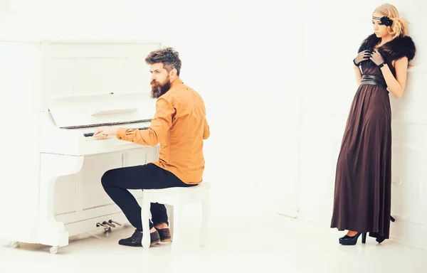 Бородатый мужчина в горчично-желтой рубашке играет на пианино в то время как красивая девушка в платье двадцатых годов и меховой воротник опирается на стену изолированы на белом фоне — стоковое фото