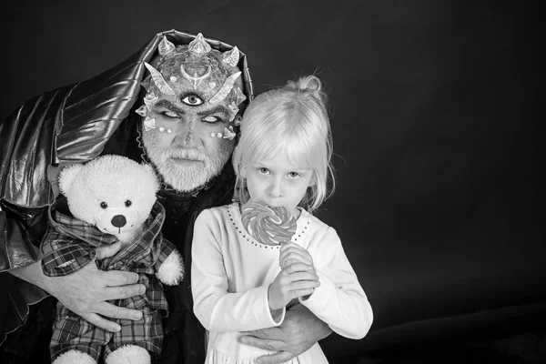 Senior man with white beard dressed like monster telling story to little girl. Fairytale concept. Man with thorns or wart with child sitting on his knees. Demon with golden hood on black background.