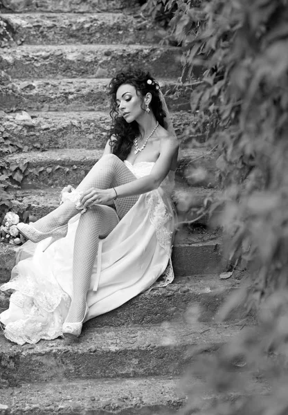 Mujer sexy en lencería medias el día de la boda. Las mujeres usan ligas de encaje en las piernas. Chica con maquillaje de novia y peinado. Novia en vestido blanco sentarse en escaleras al aire libre. Boda moda y accesorio — Foto de Stock