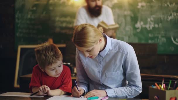 Γυναίκα με ένα αγόρι είναι ζωγραφισμένη στο τραπέζι φόντο γενειοφόρος άνδρας που διαβάζει ένα βιβλίο. Η μεθοδος διδασκαλιας. Πίσω στο σχολείο. Οι γονείς ισοπαλία με τον γιο τους σε μια σχολική τάξη. — Αρχείο Βίντεο