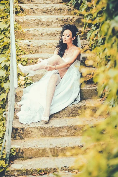 Woman wear lace garter on leg. Sexy woman in stockings lingerie on wedding day. Girl with bridal makeup and hairstyle. Bride in white dress sit on steps outdoor. Wedding fashion and accessory — Stock Photo, Image