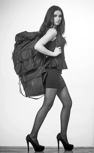 Ritratto di donna bruna in vestitino nero che si prende cura di grande zaino pesante. grazioso modello a piedi su tacchi alti isolato su sfondo bianco — Foto Stock