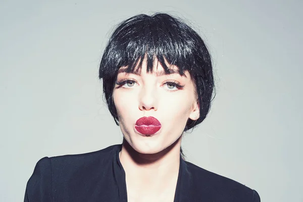 Girl on kissing face wears black formal jacket. Lady in black wig with make up on grey background. Kiss and lipstick concept. Woman with attractive red lips looks at camera.