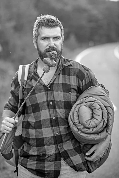 Hitchhiker on the way back to town. Bearded man with rucksack, sleeping back eating sausages from stick. Summer hiking in forest