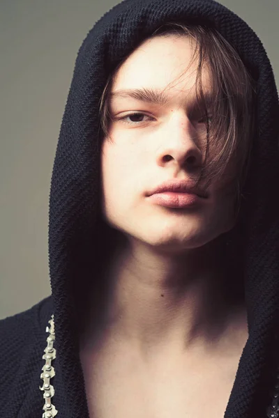 Macho in black bathrobe with hood, close up. Guy on confident face in black hood, grey background. Masculinity concept. Man with smooth skin on face and neck looking at camera.