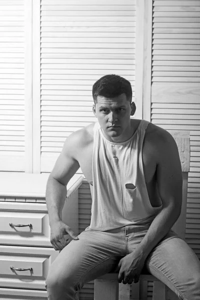 Jeune homme assis sur une chaise. Guy en débardeur tendance et jeans mode. Macho athlétique avec poitrine et mains musclées. Modèle de mode avec des cheveux élégants. Style et tendance de la mode masculine — Photo