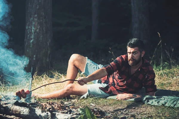 Camping and cooking food, vintage. Hipster with beard roast sausage on fire. Bearded man cook food on bonfire. Man in plaid shirt relax on nature. Tourist enjoy camping. Summer vacation concept