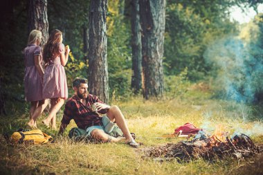 Hippi sakal oturup kamp ateşi ile. Arka plan bulanık kadın ile adam içki çay şenlik ateşi, sakallı. Arkadaşlar ormanda kamp keyfini çıkarın. Yaz tatil kavramı. Kamp ve yürüyüş ve turistik