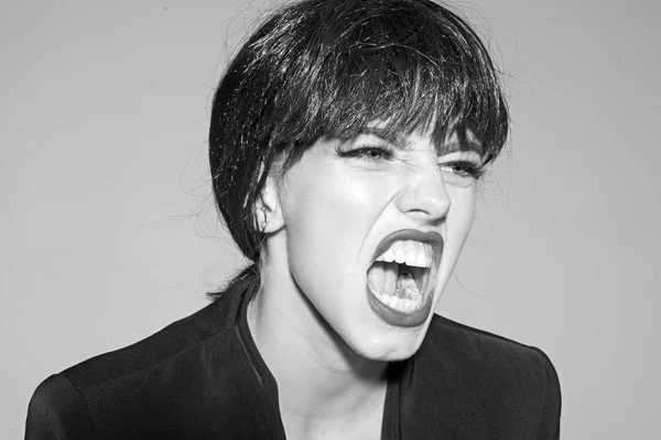 Woman with attractive red lips shouting, close up. Lady in black wig with make up screaming on grey background. Angry boss concept. Girl on scandalous shouting face wears formal jacket.