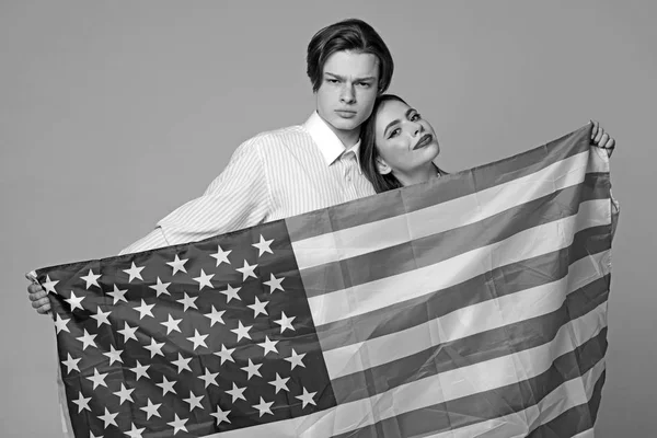 Pareja en caras tranquilas sostiene la bandera de USA. Chico y chica orgullosos de ser americanos, fondo gris. Concepto de escuela de idiomas. Pareja da la bienvenida para visitar Estados Unidos . — Foto de Stock