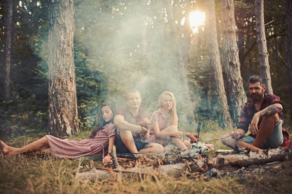 森でのキャンプの若い友人のグループです。2 つの暖かい夏の夜に楽しんでピクニックを夫婦します。キャンプファイヤー上の食糧の調理のくたびれたひげを生やした男 — ストック写真