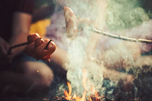 Overnachting camping in het bos. Koken worst op stokjes over vlammen voor kampvuur. Smakelijk, gerookte vlees gebakken in brand — Stockfoto