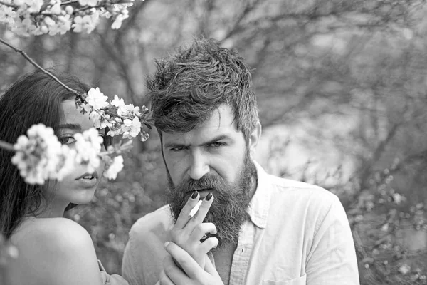 Пара курящих возле цветущих деревьев. Пара влюбленных проводят время в весеннем саду, ветви с цветами на заднем плане. Концепция курения Женские руки держат сигарету для мужчины в саду весной . — стоковое фото