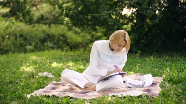 Feliz joven mujer trabajando en el portátil en el parque. Mujer sentada en el parque en la hierba verde con portátil y portátil. Encantadora estudiante sentada sobre hierba verde con cuaderno en el parque de verano . — Vídeos de Stock