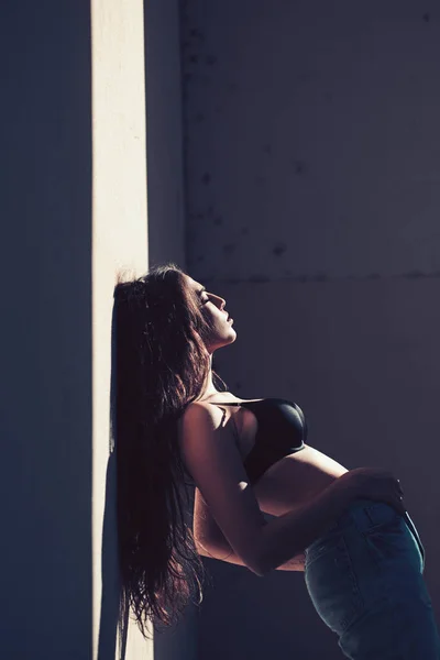 Frau mit ruhigem Gesicht lehnt an der Wand an einem sonnigen Tag. Mädchen Brünette mit nacktem Bauch entspannen unter Sonnenlicht. Dame sieht in schwarzem BH und Jeanshose attraktiv und sexy aus. heißes und sexy Konzept — Stockfoto