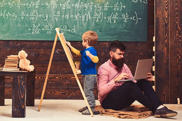 Lezione di aritmetica a scuola. Il bambino che scrive sulla lavagna mentre l'insegnante concentrato lavora sul computer portatile. Vista laterale seduto uomo e bambino in piedi back to back — Foto Stock