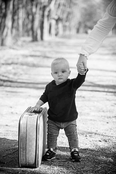 Путешествующий ребенок несет чемодан по природному ландшафту. Путешествующий мальчик путешествует с винтажной сумкой с материнской рукой — стоковое фото