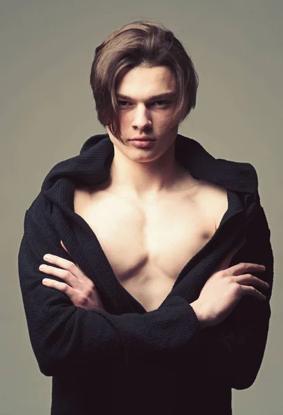 Hombre con la piel lisa en la cara, cuello y pecho mirando a la cámara. Chico con cara confiada en albornoz negro sostiene los brazos cruzados, fondo gris. Macho con corte de pelo largo con estilo. Concepto de masculinidad — Foto de Stock