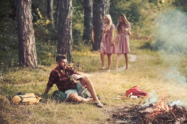 Пикник на солнечном лугу. Две девушки бродят по лесу, пока заботливый мужчина сидит на траве. Жестокий бородатый мужчина пьет кофе или чай у костра. Концепции дружбы и природы — стоковое фото