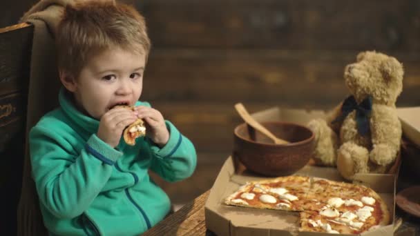 Το αγόρι τρώει πίτσα σε ένα ξύλινο υπόβαθρο. Νόστιμη πίτσα. Έχοντας μια φέτα της πίτσας αγοράκι. Πεινασμένο παιδί λαμβάνοντας μια μπουκιά από την πίτσα. Έννοια της διατροφής. — Αρχείο Βίντεο