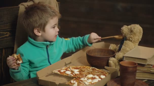 少年はピザの一部を保持し 木のスプーンからおもちゃのクマのフィードします 少年は グッズの友人とランチタイムをお楽しみください 昼食の時間 生活のため食べる ピザを食べる少年 — ストック動画