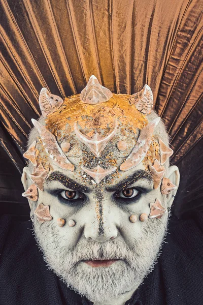 Head with thorns or warts, face covered with glitters, close up. Demon with golden collar, black background. Alien, demon, sorcerer makeup. Senior man with beard, with monster makeup. Fantasy concept.