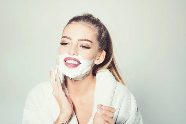 Girl on smiling face in bathrobe covering face with foam for shaving, grey background. Skin care and shaving concept. Woman with face covered with foam. Lady cares about smooth skin. — Stock Photo, Image