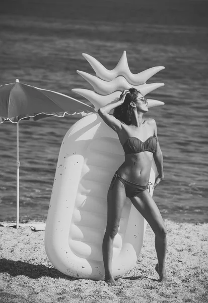 Дама с воздушным матрешкой в форме соснового яблока стоит на пляже в стильном бикини и солнцезащитных очках. Женщина держит воздушный матрас ананас в форме моря или океана на заднем плане. Летние каникулы . — стоковое фото