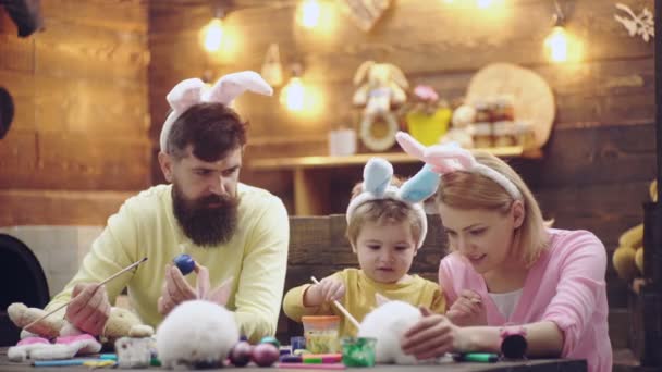 Anya, apa és fia a tojásfestés. Boldog család készül a húsvét. Aranyos kis gyermek fiú nyuszi füle visel. Húsvéti tojás fa háttér. — Stock videók